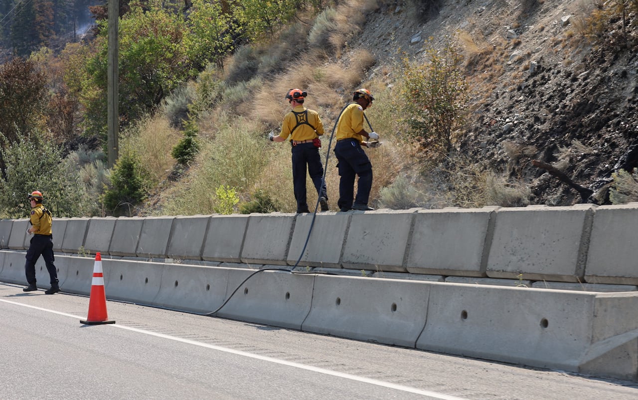 Firefighters in yellow gear seen near a highway, using hoses on blackened ground.